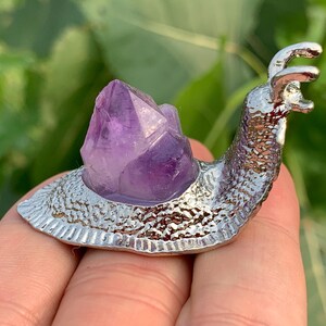 May include: A decorative snail ornament with a silver-coloured body and a large, purple amethyst crystal shell. The snail's body has a textured, ridged pattern. The crystal is a vibrant shade of purple.