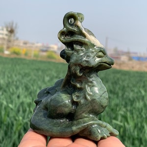 May include: A green jade sculpture of a mythical creature with a crown and intricate details. The sculpture is sitting on a hand.