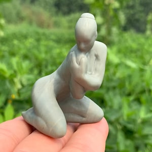 Peut inclure: Petite sculpture grise d'une personne agenouillée, les mains jointes devant elle. La figurine a une coiffure en chignon et est placée sur un fond vert flou. La sculpture est tenue dans une main.