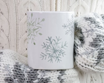 May include: White ceramic mug decorated with watercolor snowflakes in shades of green and gray. The mug is held by a person wearing a white cable-knit sweater and gray and white snowflake-patterned mittens.