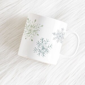May include: White ceramic mug with a curved handle, decorated with watercolor snowflakes in shades of green and blue. The mug is set against a white, fluffy background, suggesting a winter or holiday theme.