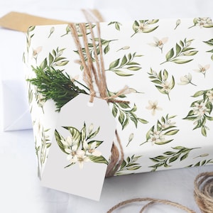 May include: A white gift box wrapped in floral patterned paper with a sprig of greenery and a white gift tag tied with twine. The paper features a repeating pattern of green leaves and white flowers.