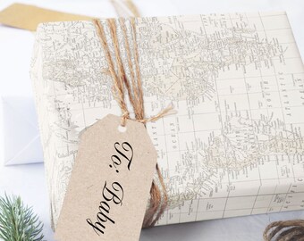 May include: A wrapped gift box with a world map design, tied with twine and a kraft paper tag that reads "To: Baby". A sprig of evergreen and a second gift box are in the background.