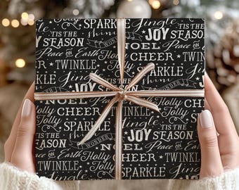 May include: A square gift box wrapped in black paper with white holiday-themed typography including words like Joy, Sparkle, Jingle, and Tis the Season. The box is tied with a thin, light-colored ribbon in a cross pattern centered on the front.