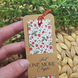 May include: A brown bookmark with a red string attached. The bookmark has a white background with a pattern of red strawberries and green leaves. The text "Just One More Chapter" is printed on the bookmark.