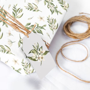 May include: A white gift box with a floral pattern of green leaves and white flowers. The box is tied with twine and has a white gift tag with a floral design.
