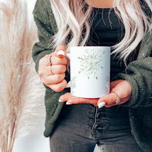 May include: A white ceramic mug featuring a watercolor snowflake design in shades of green and gray. The mug is held by a person wearing a green cardigan and dark jeans. The mug is a winter-themed item.