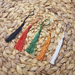 May include: Five colourful tassel ornaments with different colours, including black, red, green, orange, and white, are displayed on a woven straw surface.