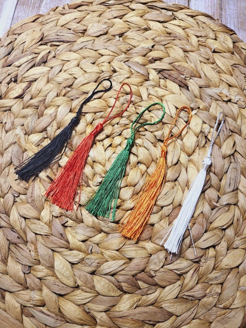 May include: Five colourful tassel ornaments with different colours, including black, red, green, orange, and white, are displayed on a woven straw surface.