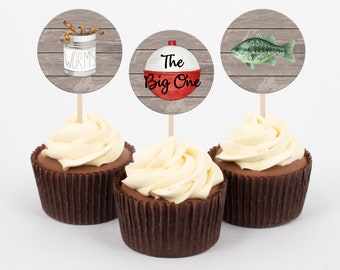 Puede incluir: Tres cupcakes con adornos temáticos de pesca. Los adornos presentan una lata de gusanos, un flotador rojo y blanco con el texto "The Big One" y un pez verde. Los cupcakes tienen bizcocho de chocolate y glaseado blanco.