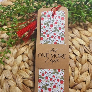 May include: A brown bookmark with a white background featuring a strawberry pattern. The bookmark has a red tassel and the text "Just One More Chapter" printed on it.