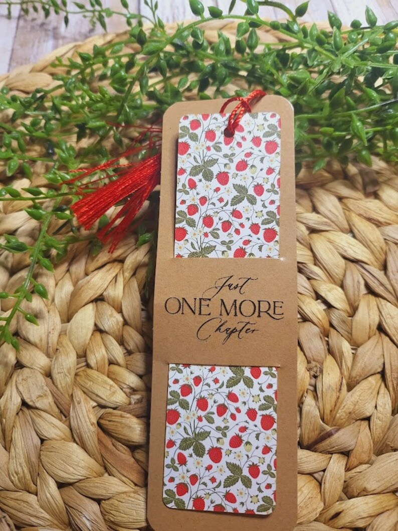 May include: A brown bookmark with a white background featuring a strawberry pattern. The bookmark has a red tassel and the text "Just One More Chapter" printed on it.