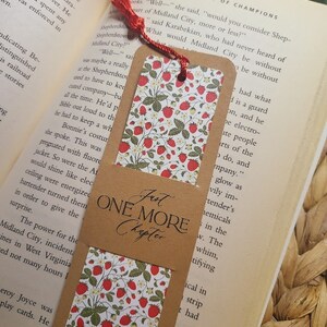 May include: A brown bookmark with a red tassel. The bookmark has a white background with a repeating pattern of red strawberries and green leaves. The bookmark has the text "Just One More Chapter" printed on it in brown.
