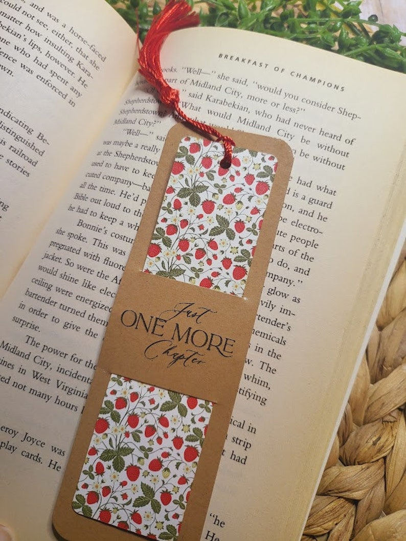 May include: A brown bookmark with a red tassel. The bookmark has a white background with a repeating pattern of red strawberries and green leaves. The bookmark has the text "Just One More Chapter" printed on it in brown.