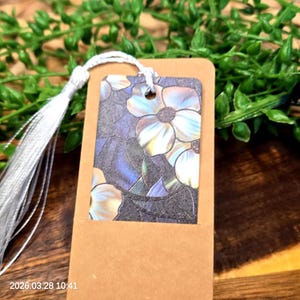 May include: A rectangular bookmark with a floral design and a silver tassel. The bookmark features a brown cardstock base with a rectangular insert showcasing a flower pattern in shades of blue, white, and iridescent colors. The tassel is attached with a white cord.