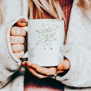 May include: White ceramic mug featuring a watercolor snowflake design in shades of green and gray. The mug is held by a person wearing a cozy, light-colored sweater. The snowflake design is repeated around the mug.