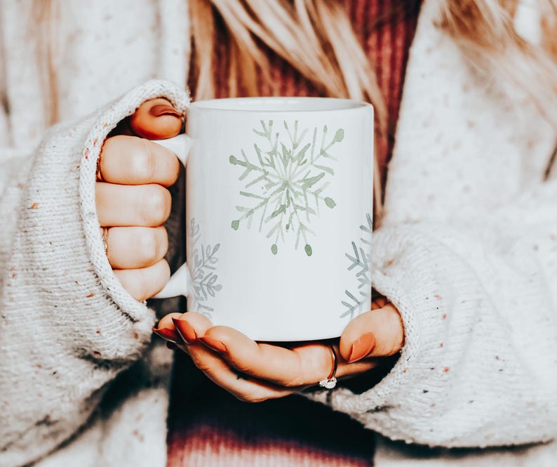May include: White ceramic mug featuring a watercolor snowflake design in shades of green and gray. The mug is held by a person wearing a cozy, light-colored sweater. The snowflake design is repeated around the mug.