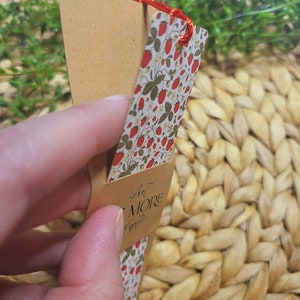 May include: A brown bookmark with a red string attached to the top. The bookmark has a floral pattern with red strawberries and green leaves on a white background. The text "Just a Little More" is written in black cursive on the bookmark.