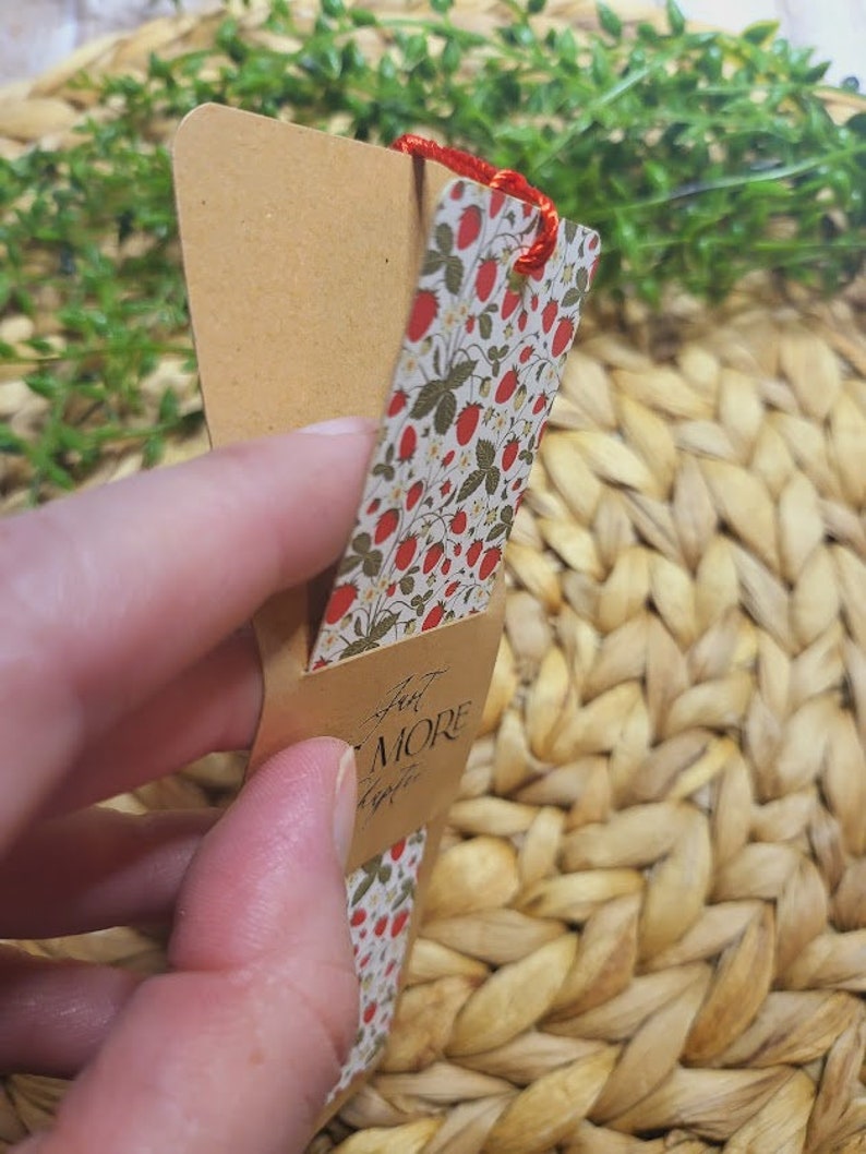 May include: A brown bookmark with a red string attached to the top. The bookmark has a floral pattern with red strawberries and green leaves on a white background. The text "Just a Little More" is written in black cursive on the bookmark.