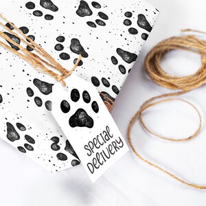 Pu&ograve; includere: Una scatola regalo bianca avvolta in carta da regalo con la stampa di impronte di zampe nere con un'etichetta regalo bianca che dice "consegna speciale". La scatola &egrave; legata con lo spago.