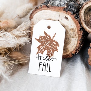 May include: A white gift tag with a brown maple leaf design and the text "Hello Fall".