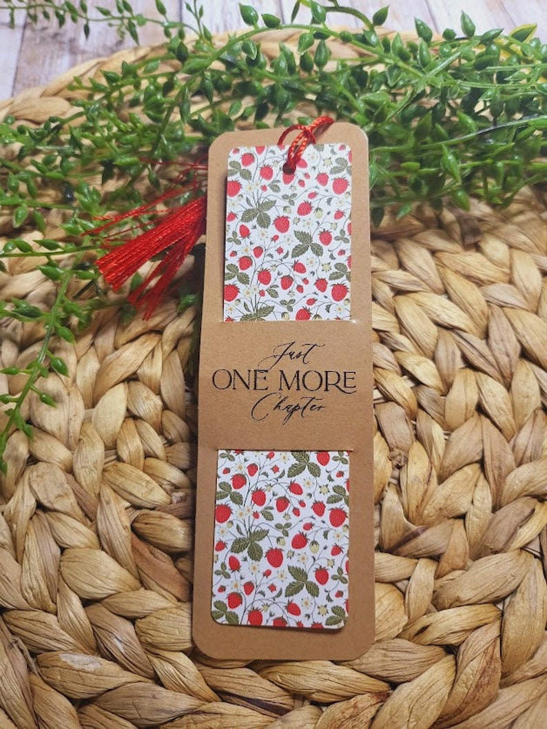 May include: A brown bookmark with a red tassel. The bookmark has a white background with a red strawberry pattern and the text "Just One More Chapter".