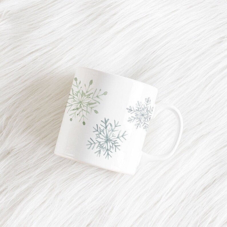 May include: White ceramic mug with a curved handle, decorated with watercolor snowflakes in shades of green and blue. The mug is set against a white, fluffy background, suggesting a winter or holiday theme.