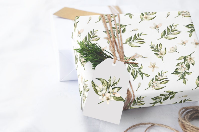 May include: A white gift box wrapped in floral patterned paper with a sprig of greenery and a white gift tag tied with twine. The paper features a repeating pattern of green leaves and white flowers.