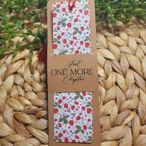 May include: A brown bookmark with a red string attached. The bookmark has a white background with a pattern of red strawberries and green leaves. The text "Just One More Chapter" is printed on the bookmark in black.