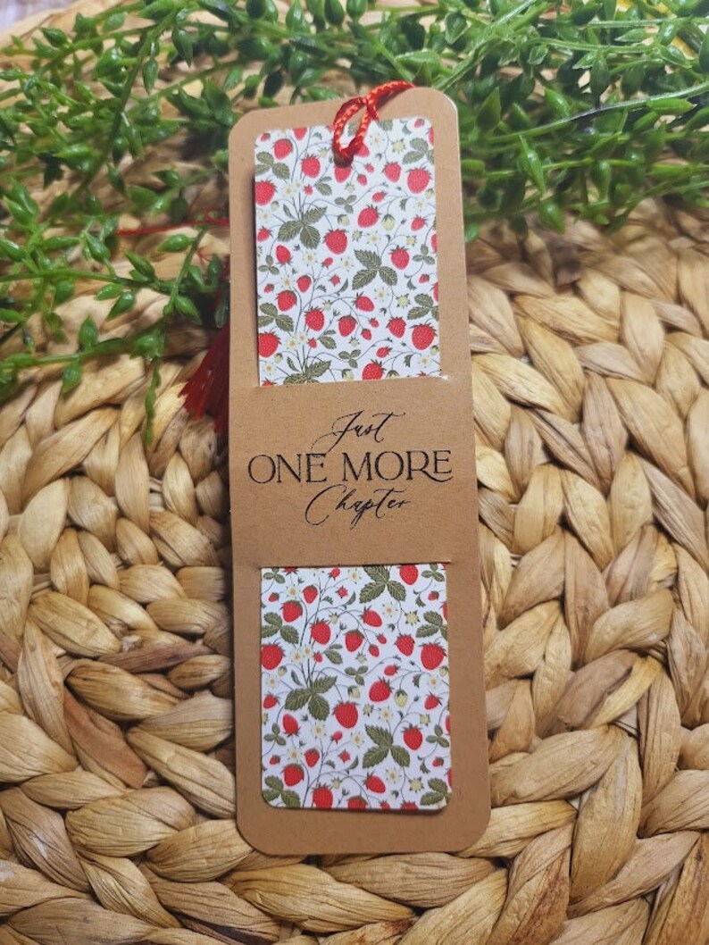 May include: A brown bookmark with a red string attached. The bookmark has a white background with a pattern of red strawberries and green leaves. The text "Just One More Chapter" is printed on the bookmark in black.