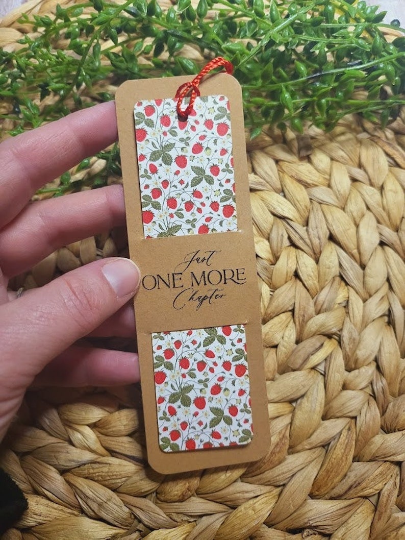 May include: A brown bookmark with a red string attached. The bookmark has a white background with a pattern of red strawberries and green leaves. The text "Just One More Chapter" is printed on the bookmark.