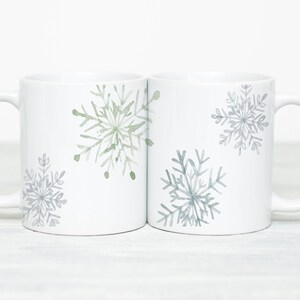 May include: Two white ceramic mugs with snowflake designs. Each mug features a large snowflake in shades of gray and green. The mugs have a simple, elegant design, perfect for winter or holiday gifting.