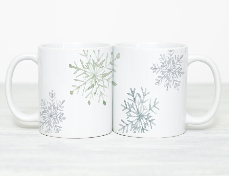 May include: Two white ceramic mugs with snowflake designs. Each mug features a large snowflake in shades of gray and green. The mugs have a simple, elegant design, perfect for winter or holiday gifting.