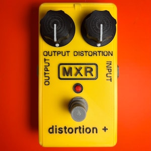 May include: A yellow MXR distortion guitar effects pedal with black knobs and the text "OUTPUT DISTORTION" and "INPUT". The pedal has a red indicator light and the text "distortion +" on the bottom. The background is a solid orange.