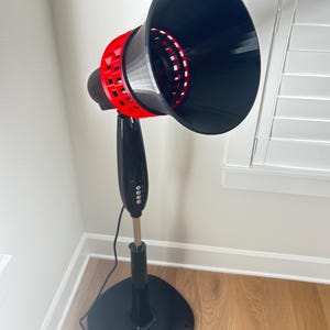 May include: A black and red electric fan with a round base and a tall, adjustable stand. The fan head is large and cone-shaped, with a red, perforated ring around the center. The fan is positioned in front of a white wall and a window with white blinds.