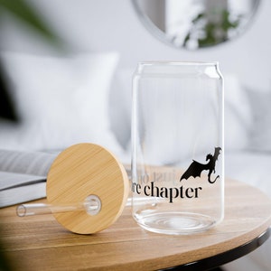 Peut inclure: Une canette en verre transparente avec un couvercle en bambou et une paille. La canette a une silhouette noire de dragon et le texte "Just one more chapter".