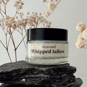 May include: A small white jar with a black lid and a label that reads "Homemade Whipped Tallow" with the ingredients listed below. The jar is sitting on a gray rock with white flowers in the background.