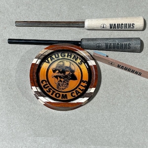 May include: A wooden circular coaster with a brown, orange, and white striped design. The center of the coaster features a logo with a cartoon turkey wearing sunglasses and a hat. The logo reads "Vaughn's Custom Calls".