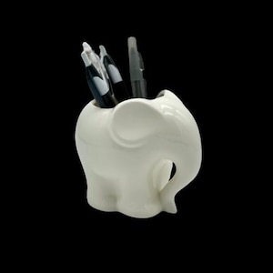 Może przedstawiać: Biały ceramiczny organizer na biurko w kształcie słonia z trzema czarnymi długopisami w środku.