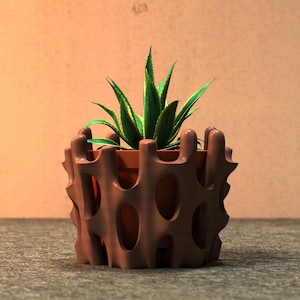 May include: A brown, decorative plant pot with a unique, openwork design. The pot holds a small succulent plant with green and yellow leaves. The pot is sitting on a gray surface, with a light brown background.