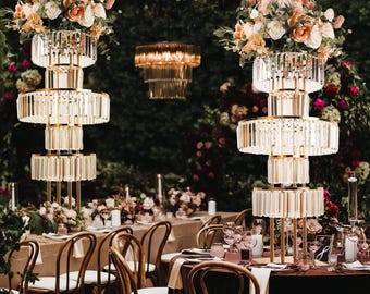 May include: Two gold metal tiered stands with crystal accents, decorated with pink and white flowers, are used as centerpieces for a wedding reception. The stands are placed on a table with brown linens and surrounded by chairs.