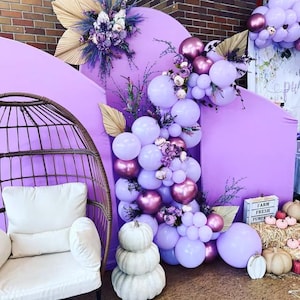 May include: A purple backdrop with a balloon arch made of lavender and metallic burgundy balloons. The arch is decorated with greenery and flowers. A white wicker chair sits in front of the backdrop. A sign reads "Farm Fresh Pumpkins".
