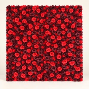 May include: A square wall panel densely covered in a mix of artificial flowers, featuring a vibrant color palette of bright red, deep crimson, and dark burgundy roses and smaller filler blossoms arranged in a uniform, textured floral display.