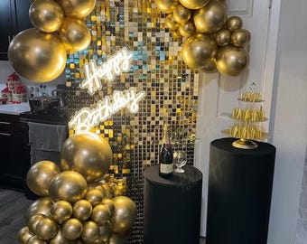 Könnte beinhalten: Goldener Ballonbogen mit goldfarbenem Paillettenhintergrund und einem Neon-Schild mit der Aufschrift "Happy Birthday". Es gibt zwei mit Schwarz bezogene Tische mit einer Flasche Champagner und einem Glas auf dem einen Tisch und einem mehrstöckigen Kuchenständer auf dem anderen.