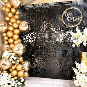 Pode incluir: Um fundo preto de lantejoulas com uma placa circular dourada que diz "Be Mine". Há balões dourados e flores brancas de cada lado do fundo.