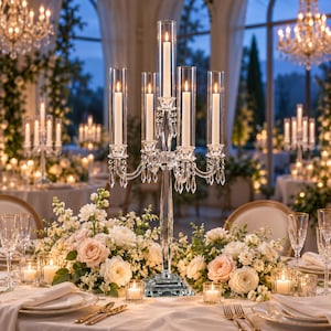 May include: A tall, clear glass candelabra featuring five candle holders with cylindrical glass sleeves, accented by hanging crystal prisms. The structure stands on a square base, holding five lit white taper candles in a formal setting.