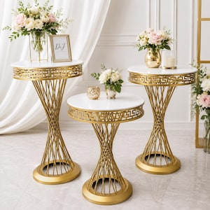 May include: Three round pedestal tables with white circular tops and gold-colored metal bases featuring a geometric Greek key pattern along the rim and a twisted wire frame design. Each table stands on a solid circular gold-colored metal base.