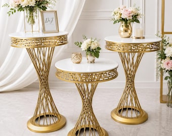 May include: Three round pedestal tables with white circular tops and gold-colored metal bases featuring a geometric Greek key pattern along the rim and a twisted wire frame design. Each table stands on a solid circular gold-colored metal base.