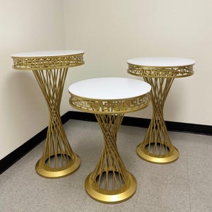 May include: Three gold metal cake stands with white marble tops. The stands have a geometric design around the top edge and a twisted wire base.