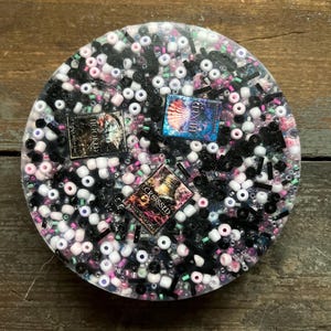 May include: A round, clear resin coaster with black, white, pink, and green beads embedded in the resin. There are three book covers embedded in the resin, one with the title "HEXED", one with the title "CROSSED", and one with the title "MOONLAND".